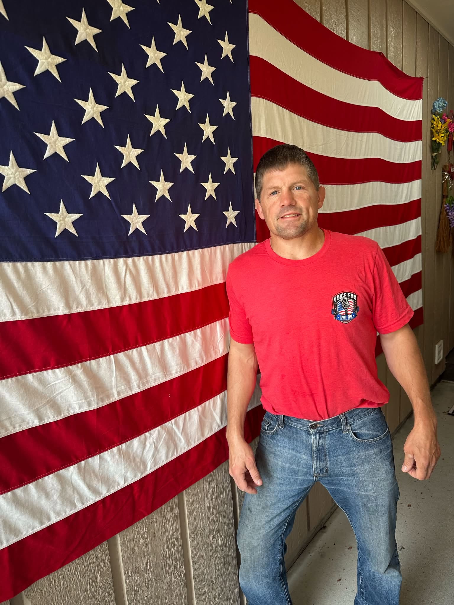 Kirk White wearing a red Voice for Valor T-shirt in front of an American flag