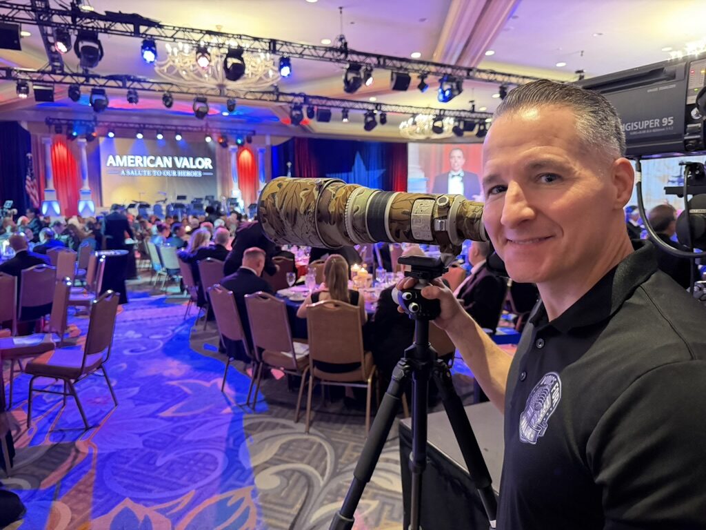 Michael Komorous behind a telephoto lens at the American Valor gala, with the 'American Valor — A Salute to Our Heroes' stage behind him.