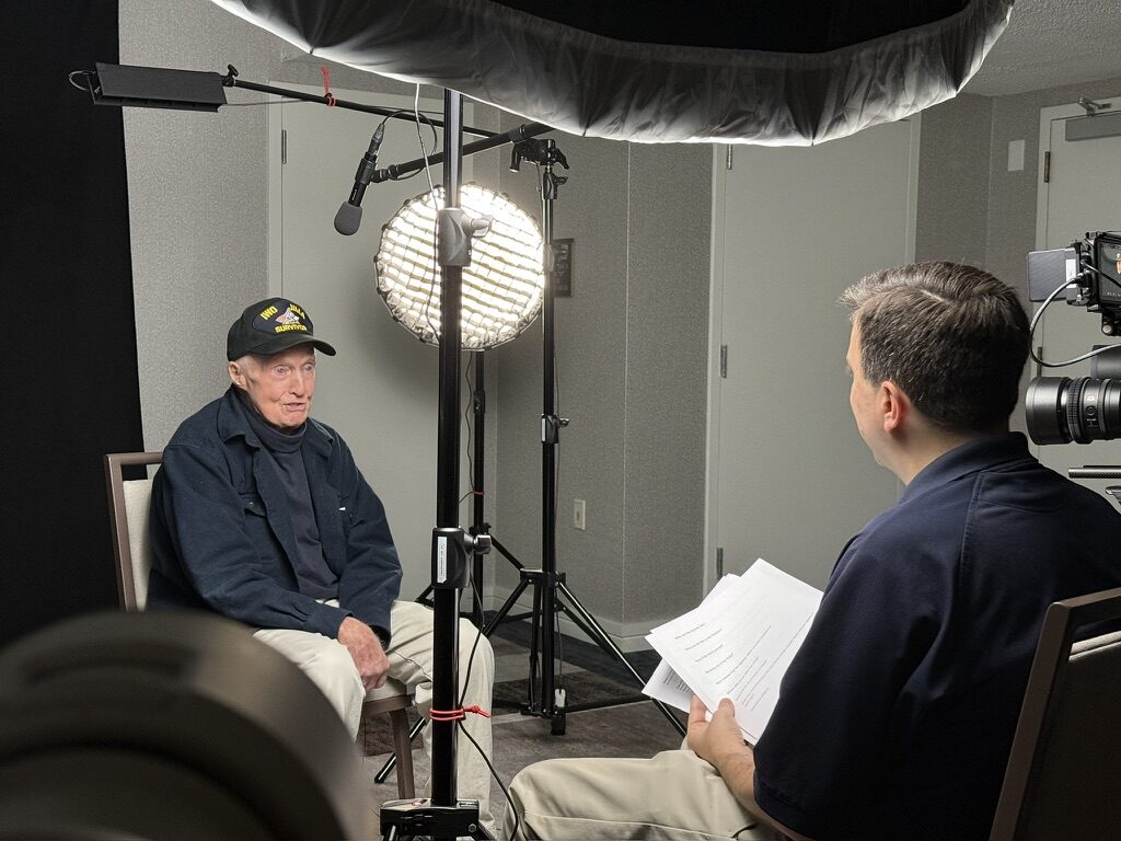 An Iwo Jima survivor, wearing a survivor's cap, sits for an oral-history interview under studio lighting.