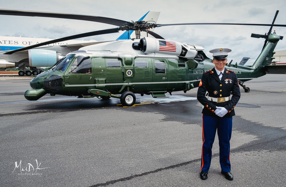 A U.S. Marine in dress blues stands guard before Marine One on the ramp in Charlotte, with Air Force One behind.